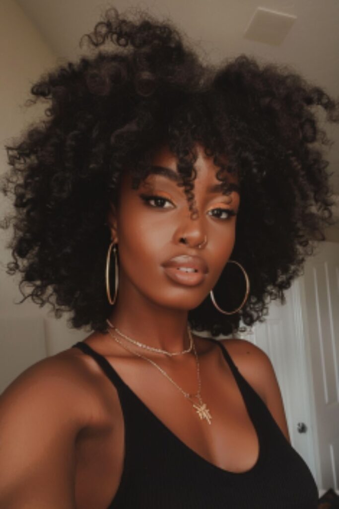A black woman with big curly afro, wearing a black top, a simple necklace with a charm on it, taking a mirror selfie of her profile in her bedroom, has beautiful makeup. She has large lips and full eyelashes, she's smiling. Her hair is shoulder length in black afro.