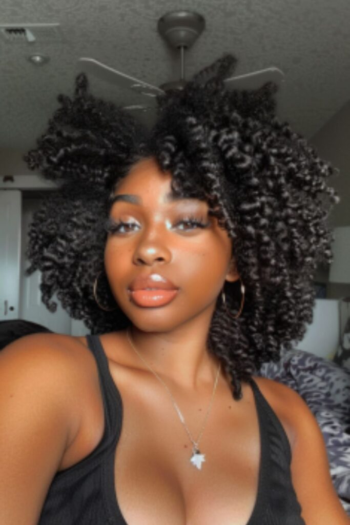 This image captures a woman with a lush, curly crochet hairstyle featuring natural brown tones. Her makeup is beautifully applied, emphasizing soft, smoky eyes, perfectly shaped eyebrows, and glossy, nude lips. She is wearing a simple black tank top and layered silver necklaces, one with a prominent pendant, adding a touch of sophistication to her casual yet polished appearance.